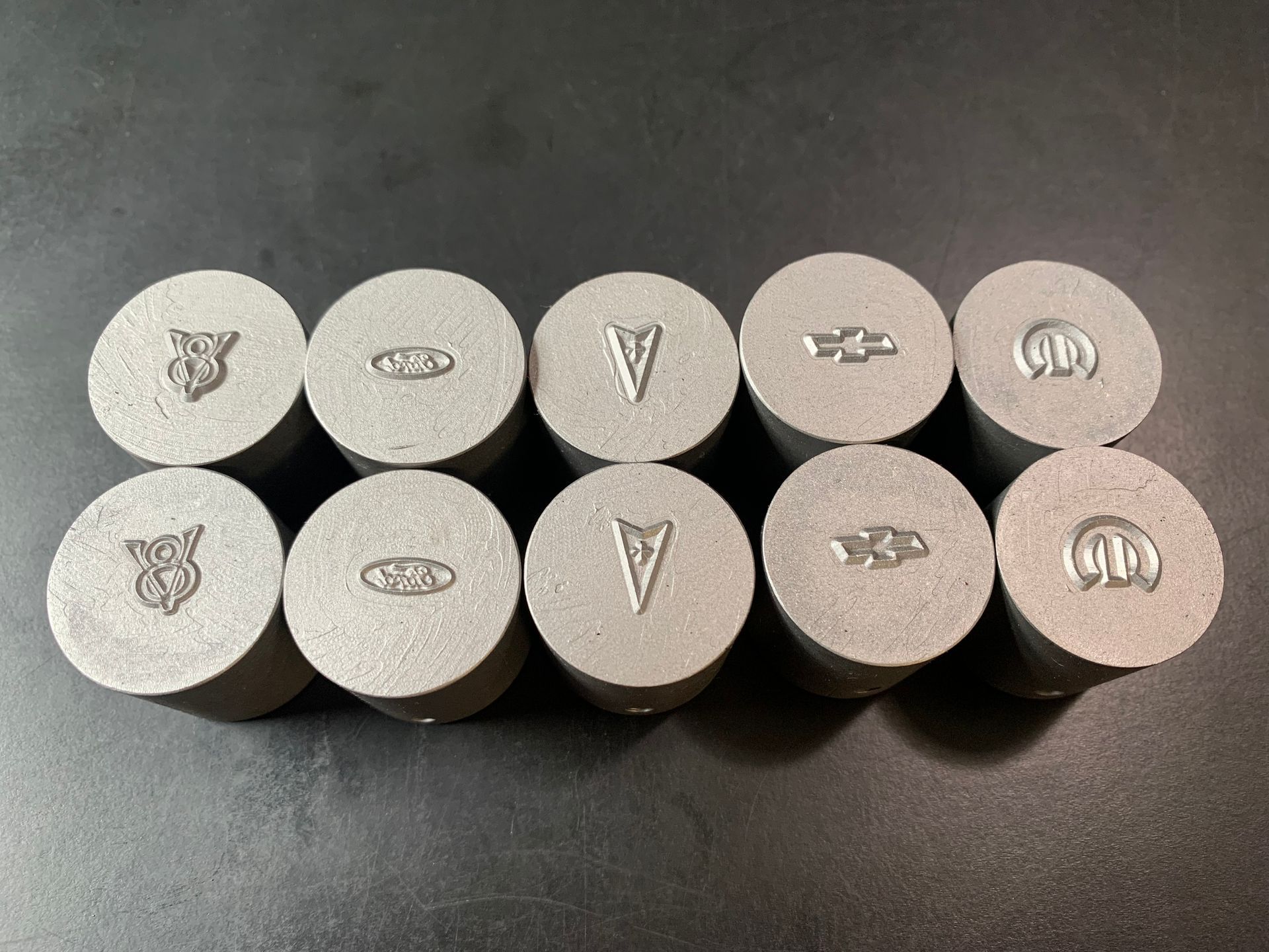 Ten metal automotive emblems arranged in two rows. Logos include Buick, Oldsmobile, Pontiac, Chevrolet, and Mercury.