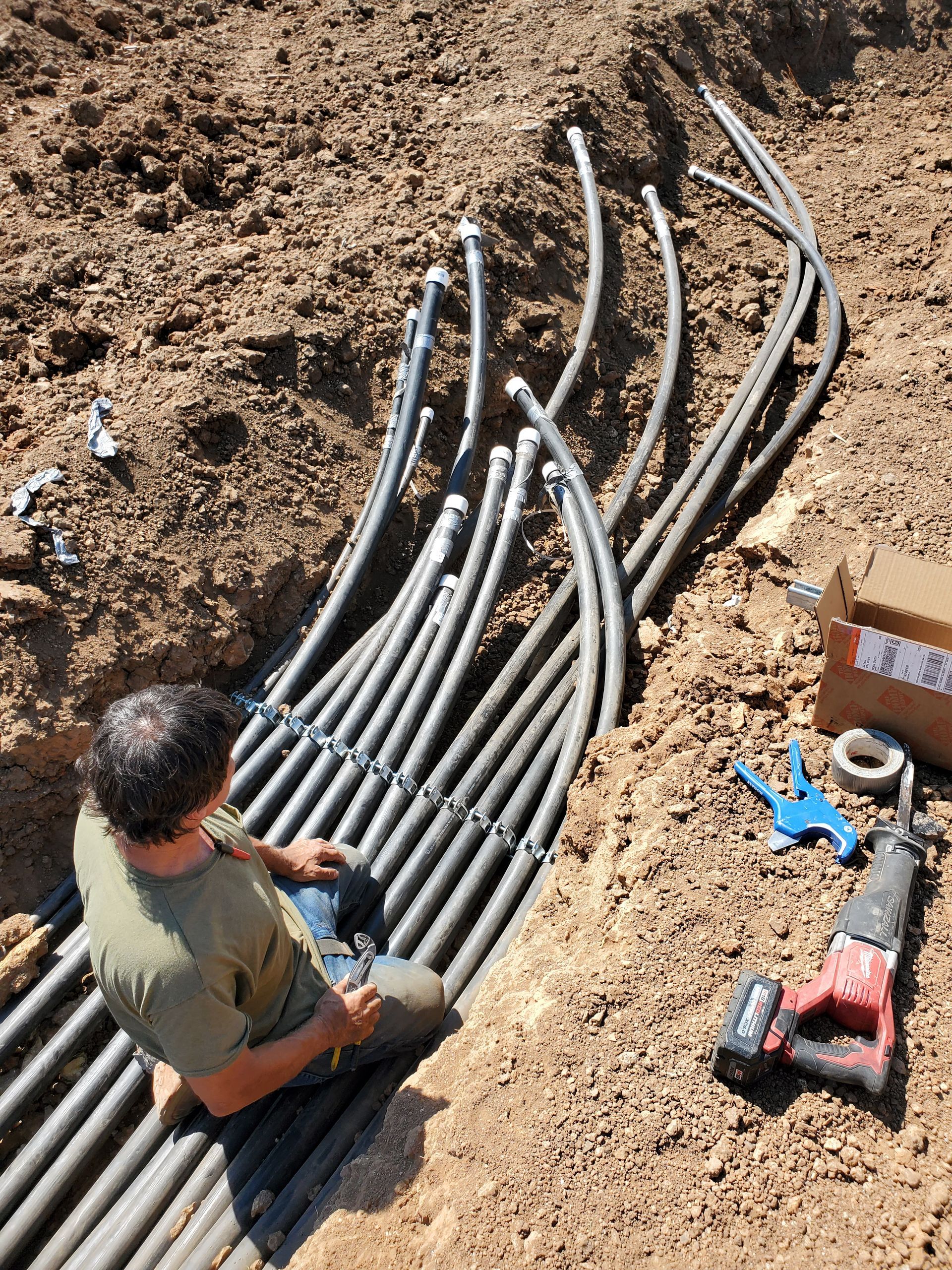 A man is sitting in the dirt working on a bunch of pipes.