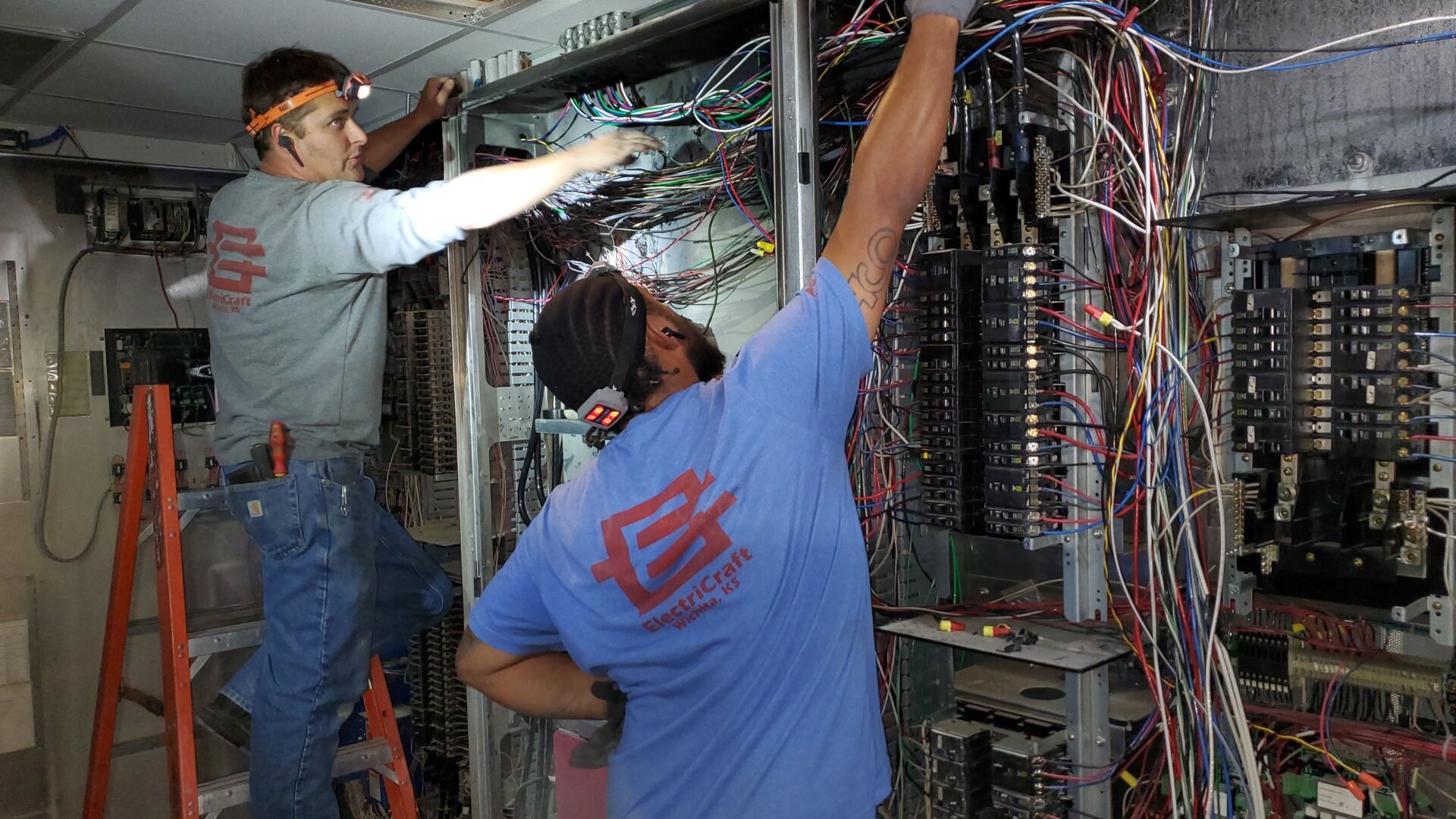 electricians working on a complex network of wires inside a cabinet. One on a ladder.
