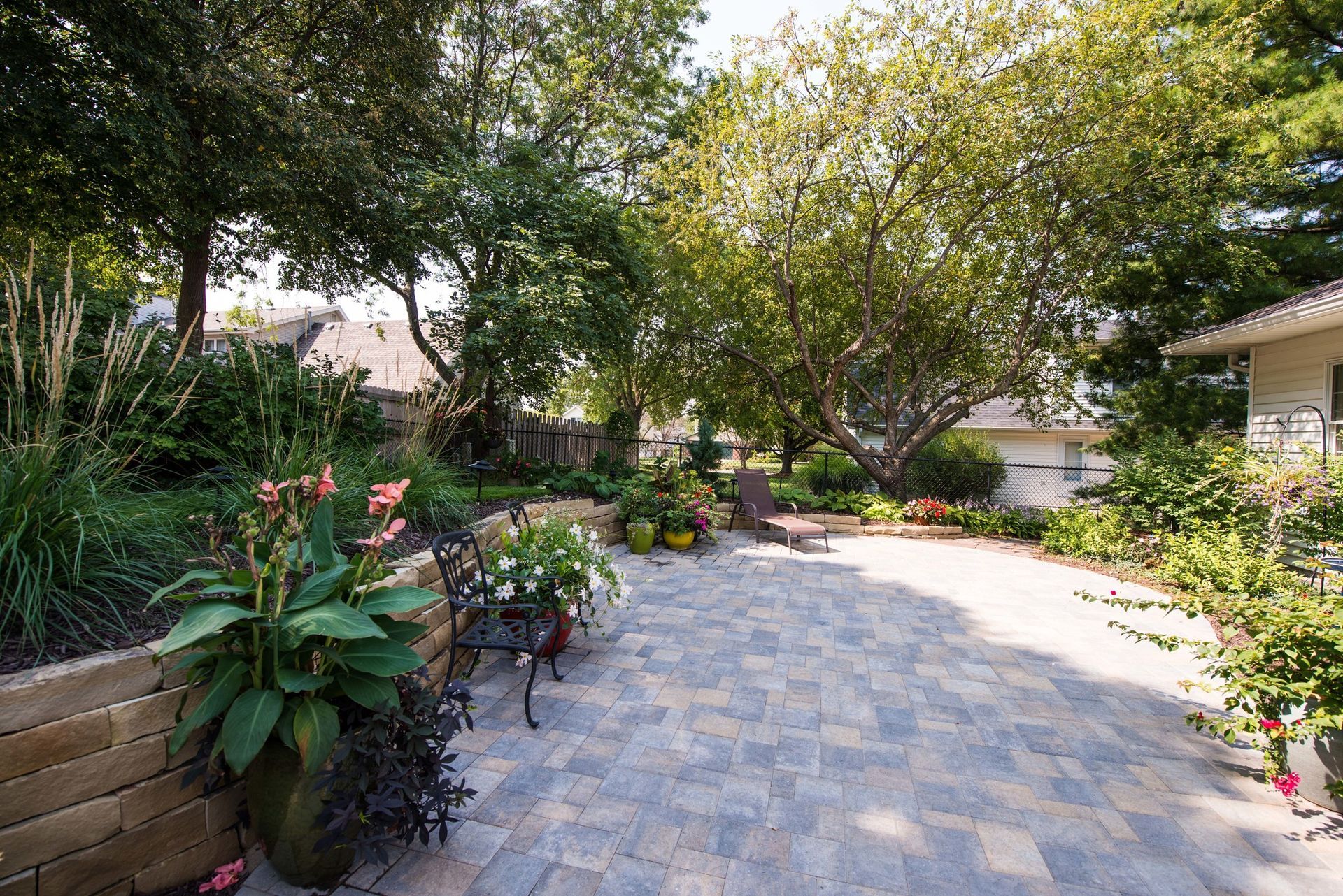 Sunlit garden patio with stone paving, potted plants, trees, and a house at right