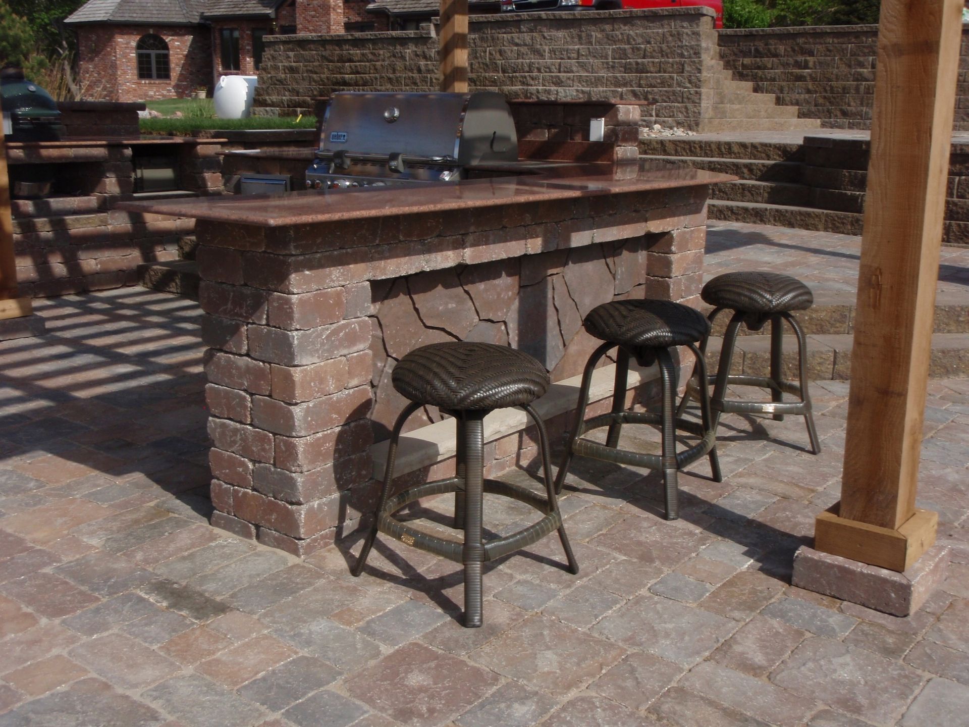 Outdoor brick patio bar with three stools and a built-in grill area in sunlight