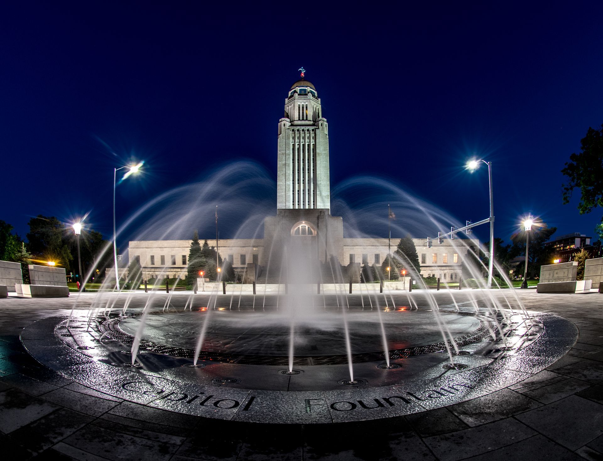 Nighttime city fountain with arcing water in front of a lit capitol building and tower