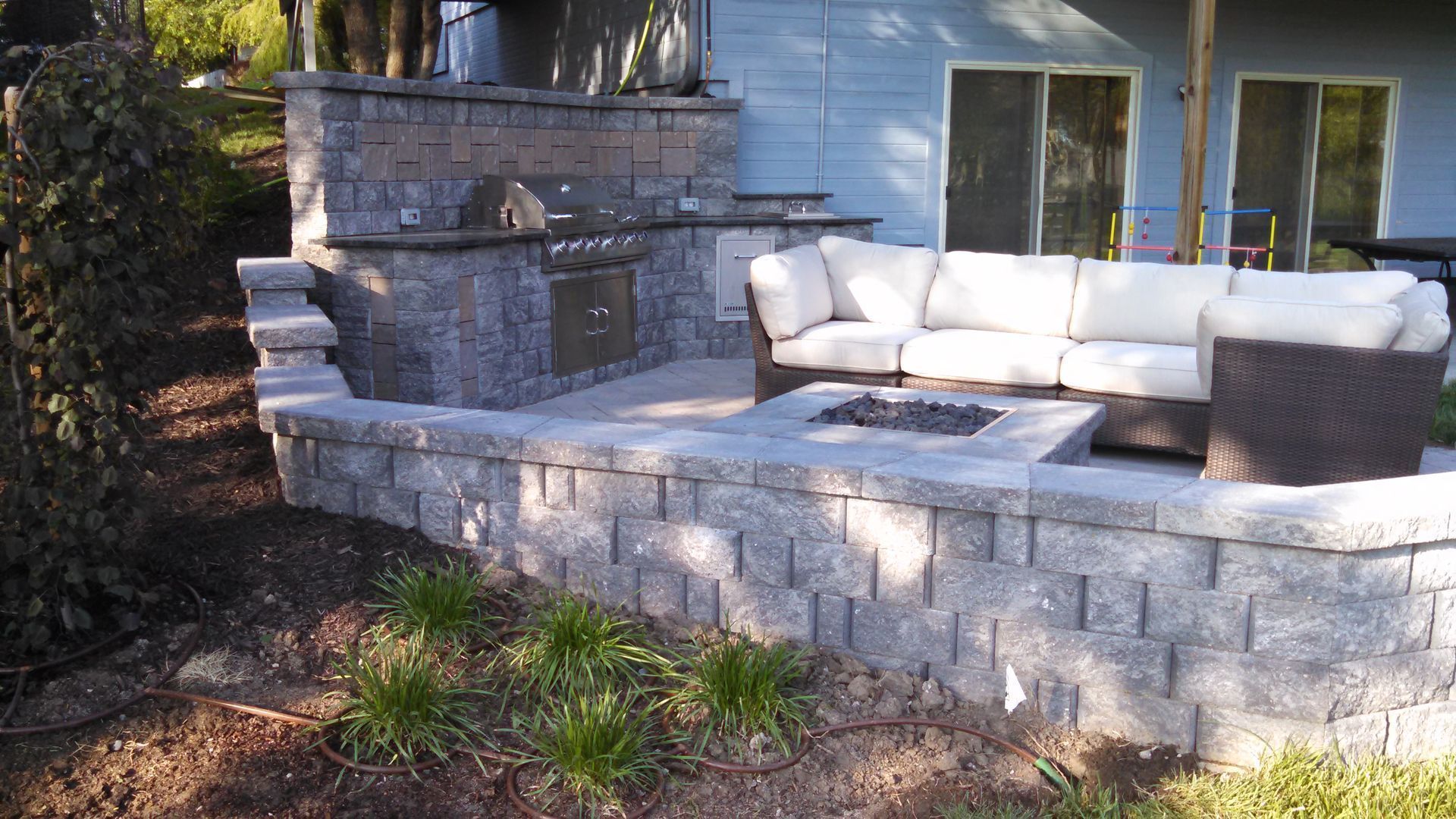 Stone patio with white outdoor sectional sofa beside a house, surrounded by trees and garden beds.