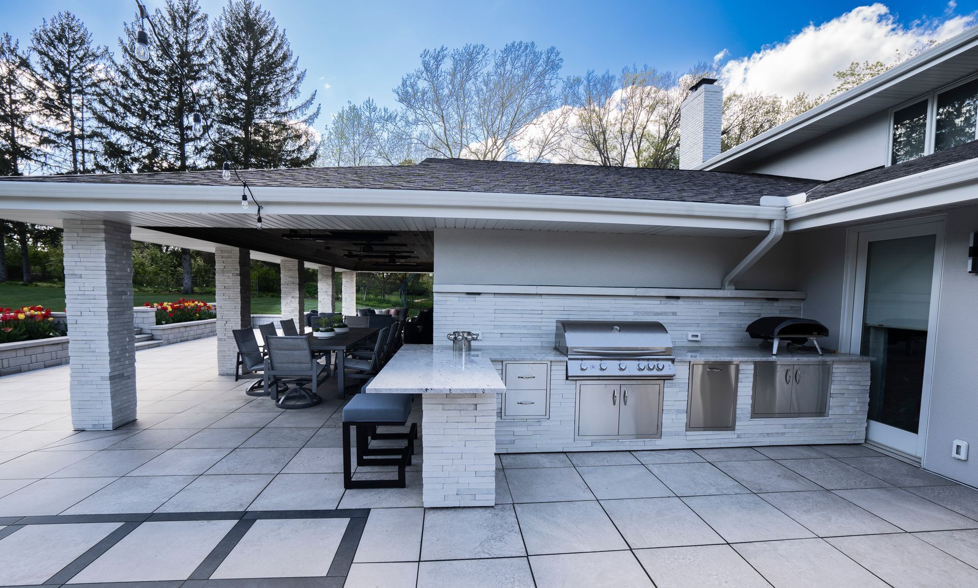 Covered outdoor patio with white stone flooring, built-in grill, sink, and seating under a white pergola.