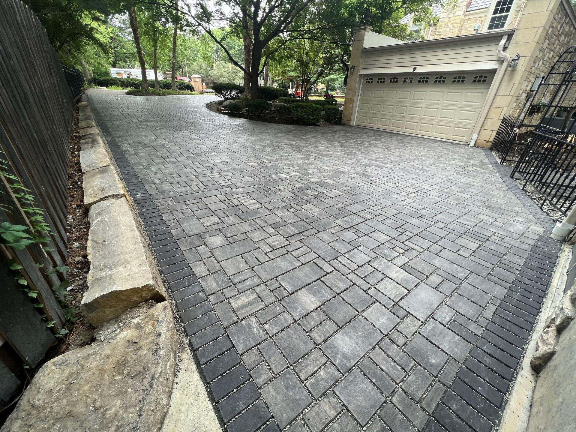 Brick driveway leading to a garage, bordered by trees and stone walls in a residential courtyard