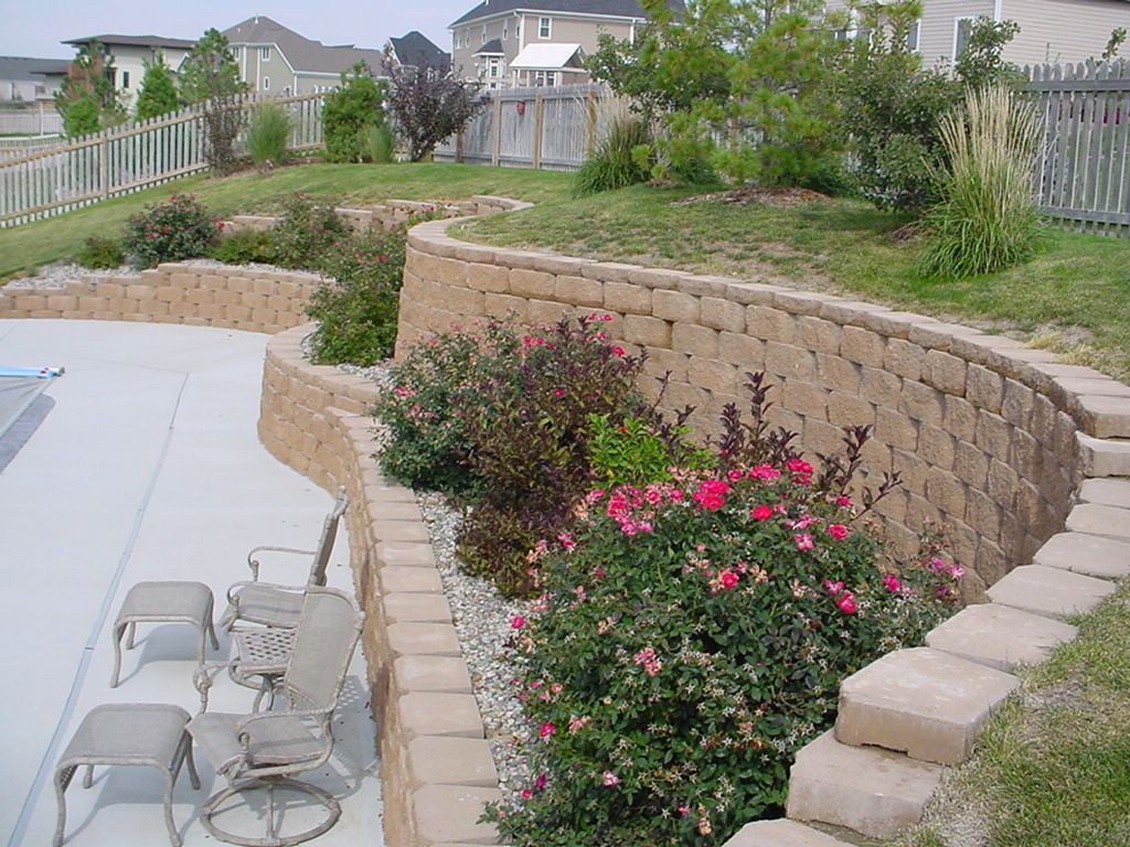 Backyard patio with white chairs, stone retaining wall, and pink flowering shrubs along a grassy slope
