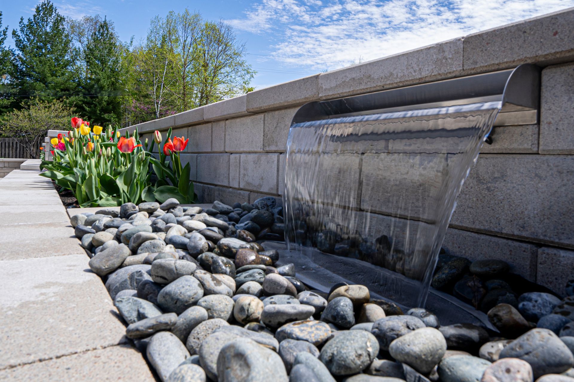 Decorative garden water feature with flowing water, river rocks, and red-orange tulips beside a stone wall