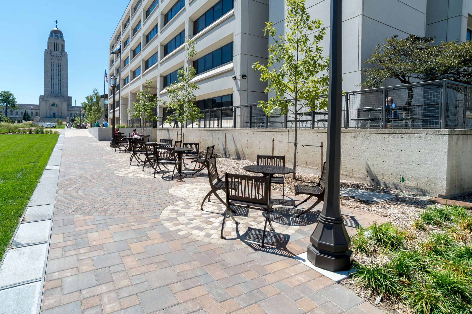Sunny sidewalk café tables beside a modern building, with a tall tower in the distance