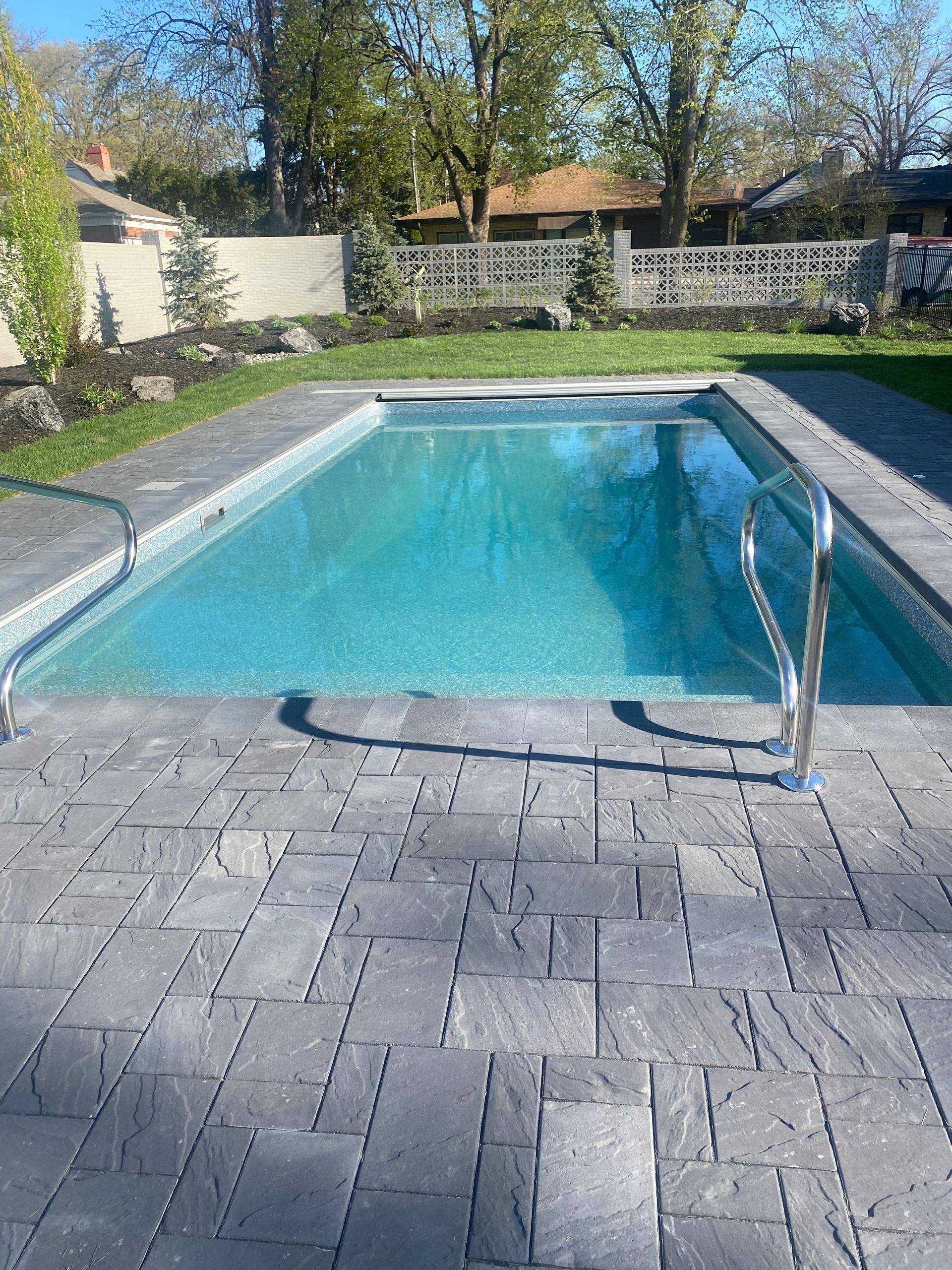 Backyard swimming pool with blue water, stone patio, and metal handrail under trees