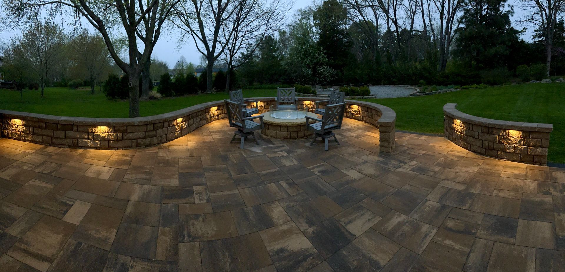 Stone patio with lit retaining walls and outdoor tables at dusk, surrounded by trees and garden