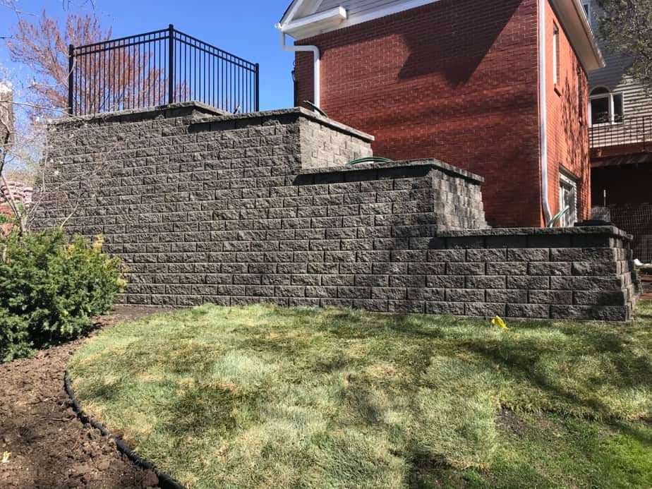 Stone retaining wall beside a red house with a black metal fence on a grassy slope.