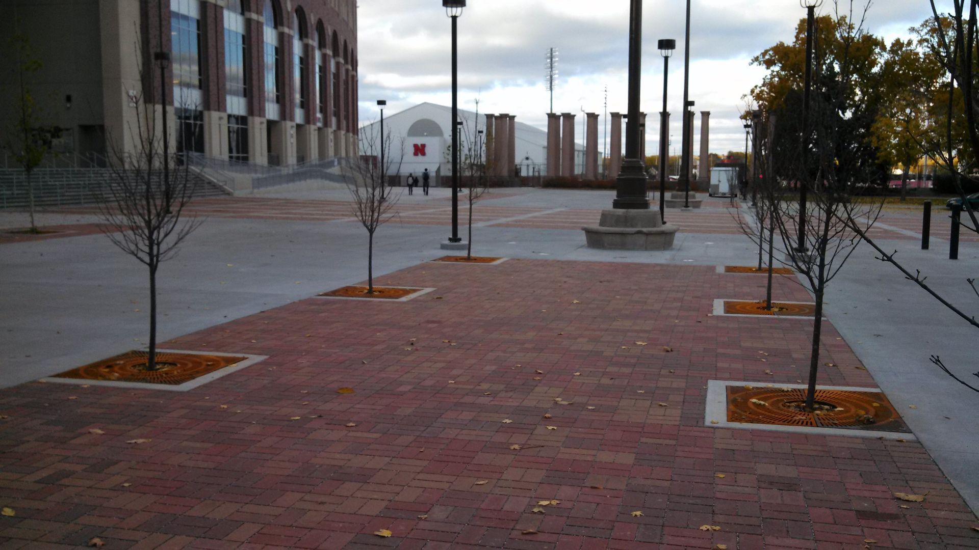 Brick-paved plaza with young trees, lamp posts, and a building walkway at dusk
