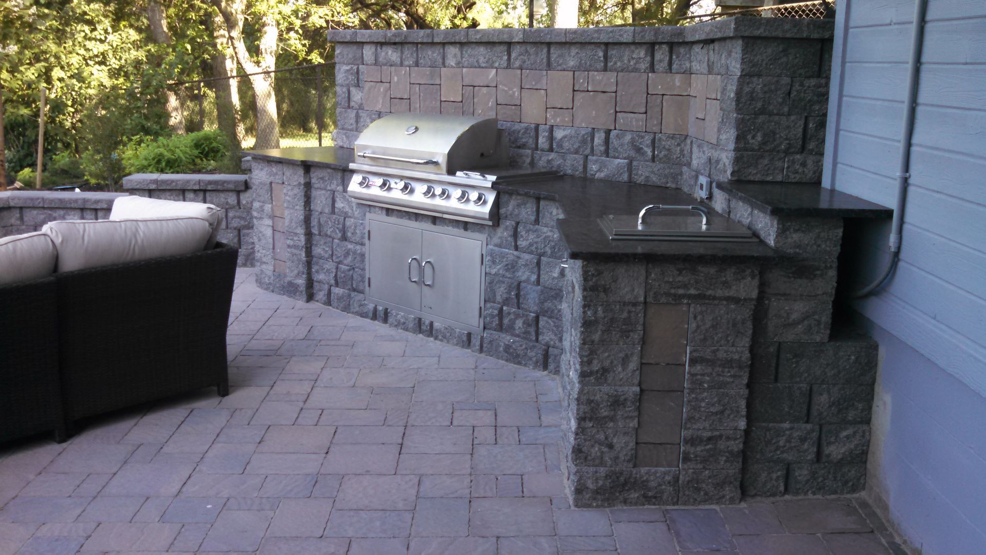 Outdoor patio with stone grill and built-in counter beside a wicker sofa and paver flooring