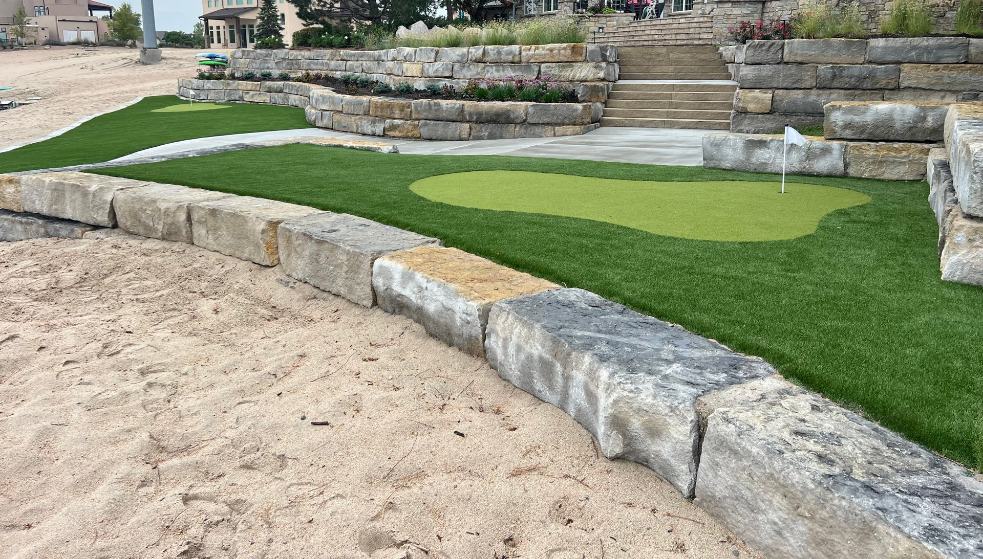 Landscaped garden with stone retaining walls and a curved patch of green artificial turf beside sand
