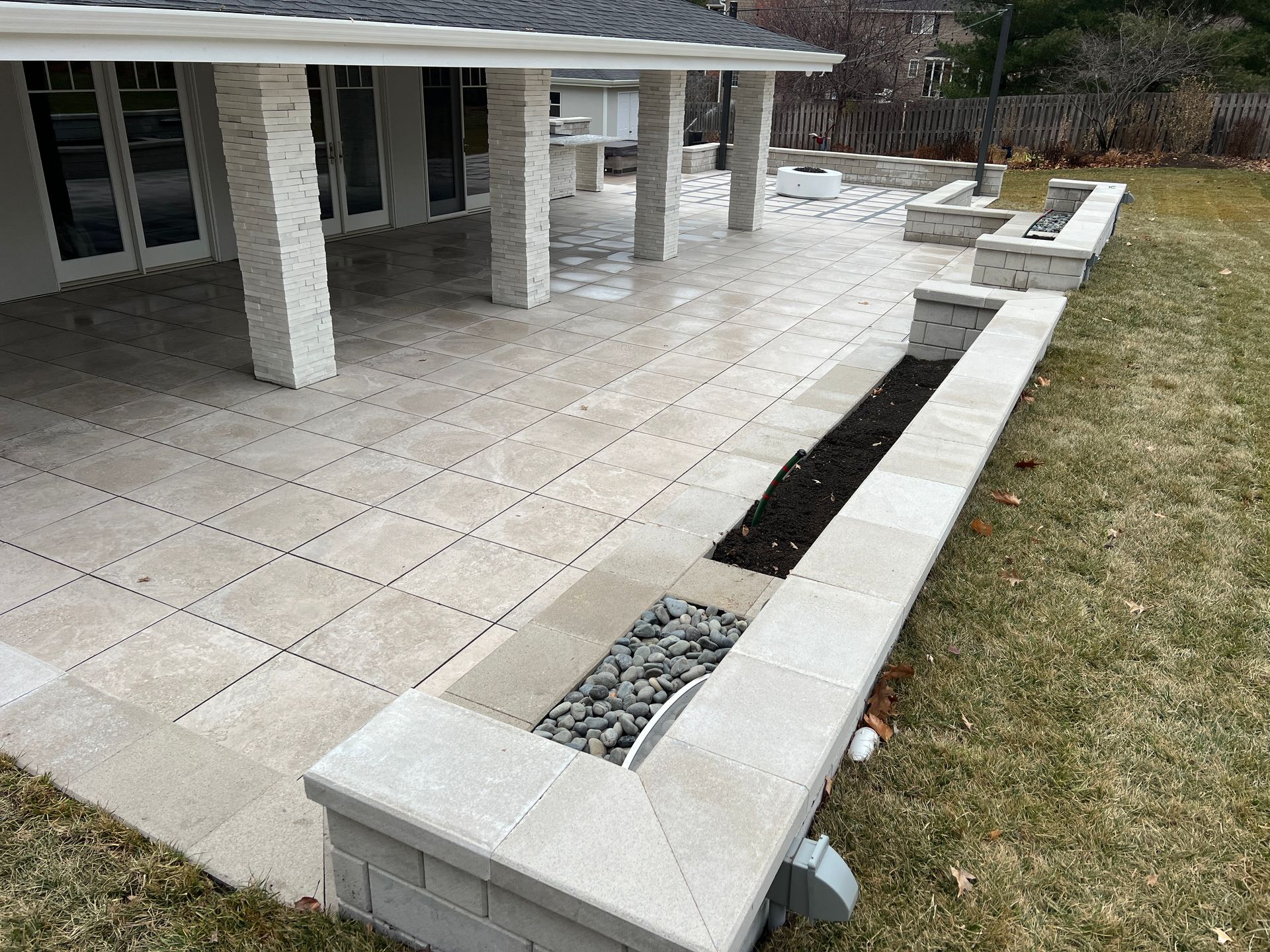 Stone patio and low planter wall beside a house, with white columns and a grassy yard.