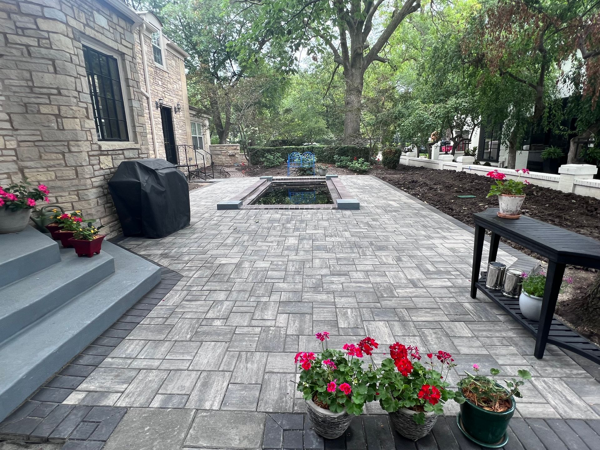 Stone patio with potted red flowers, outdoor seating, and a central fire pit beside a stone house.