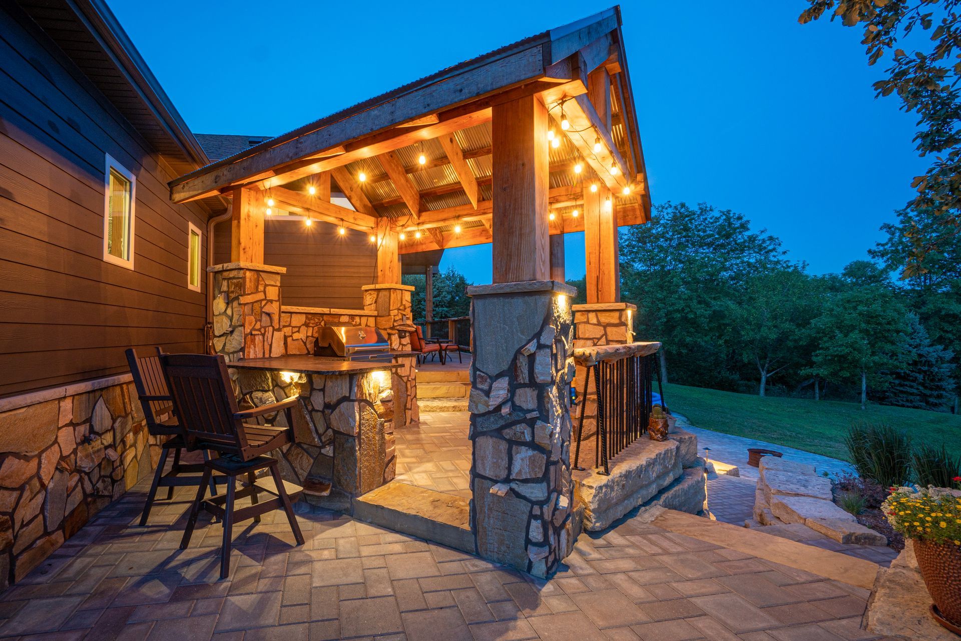 Warmly lit wooden patio with stone pillars and seating beside a house at dusk