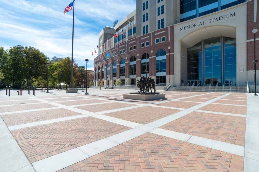 Empty plaza outside a brick stadium with a U.S. flag and blue glass entrance under a clear sky