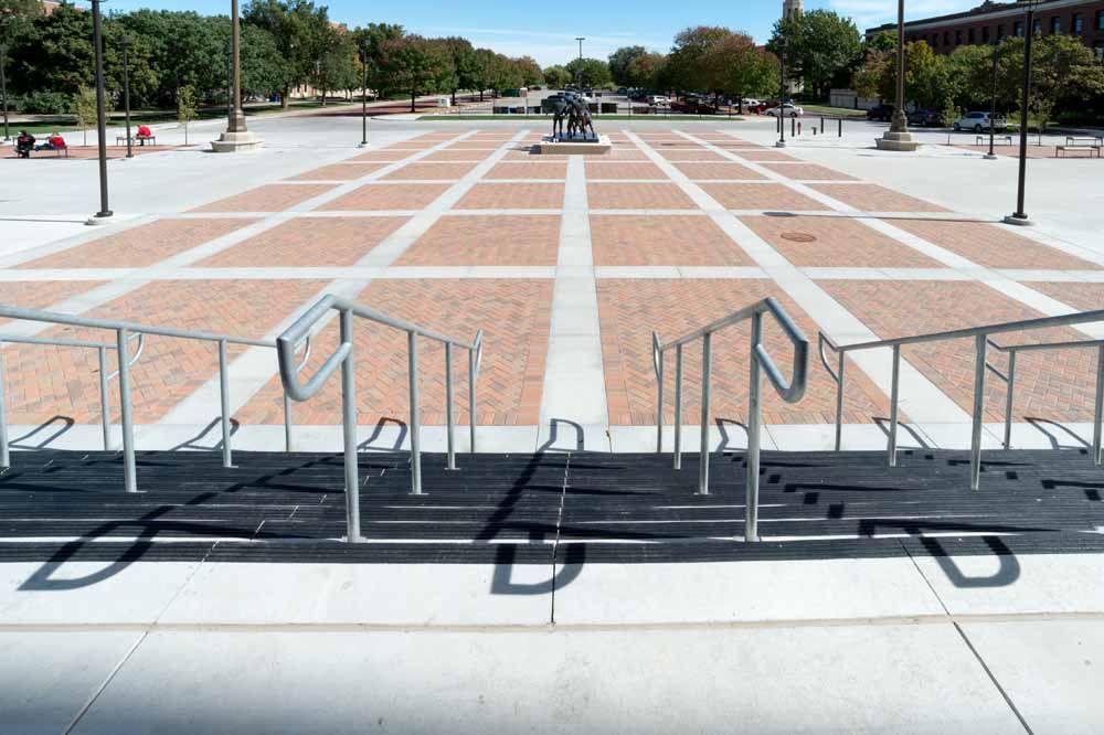 Outdoor plaza with brick paving, steps, handrails, and ramps leading to a central fountain.