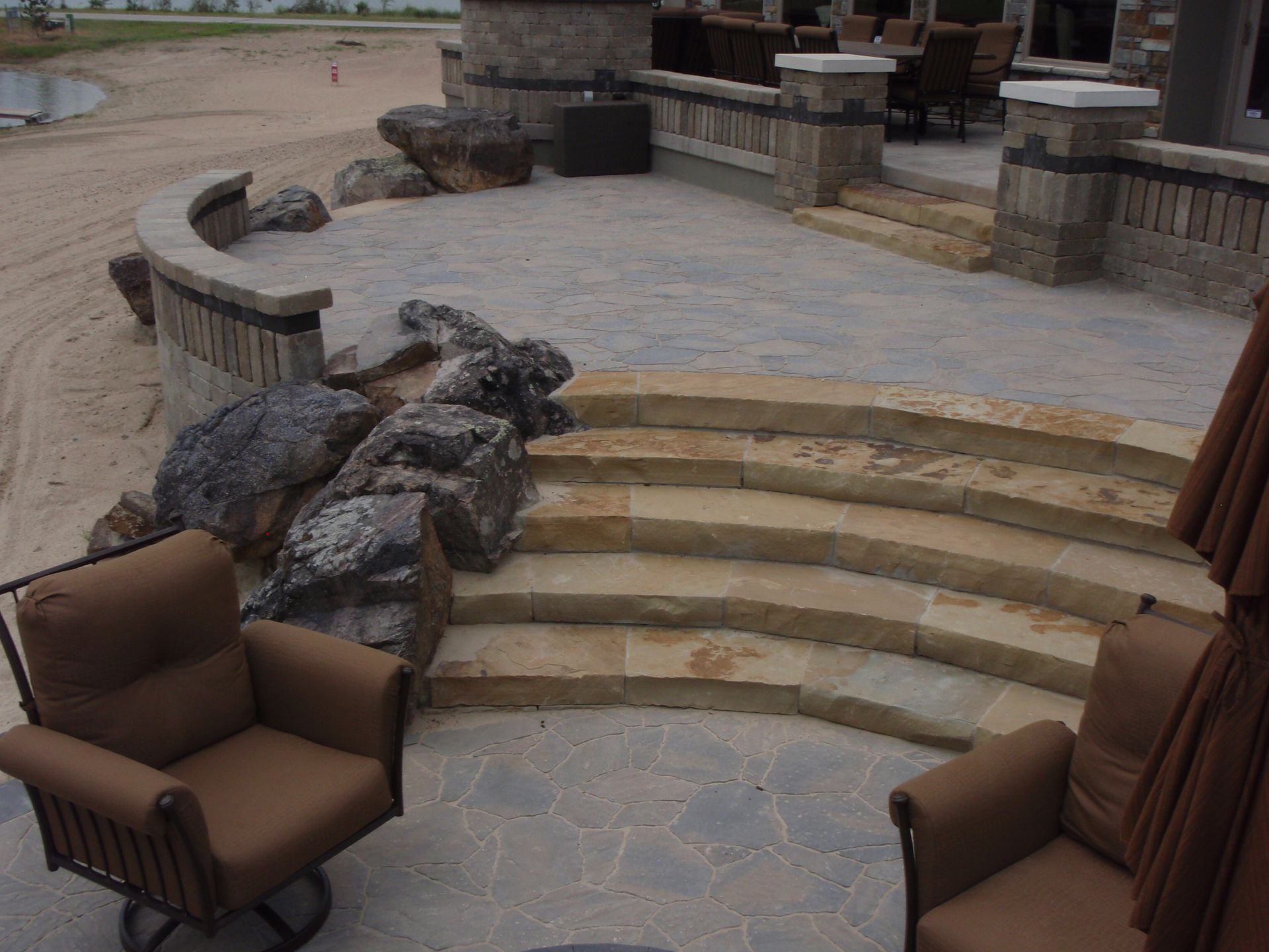 Stone patio with curved steps, built-in benches, and brown outdoor chairs around a fire pit area