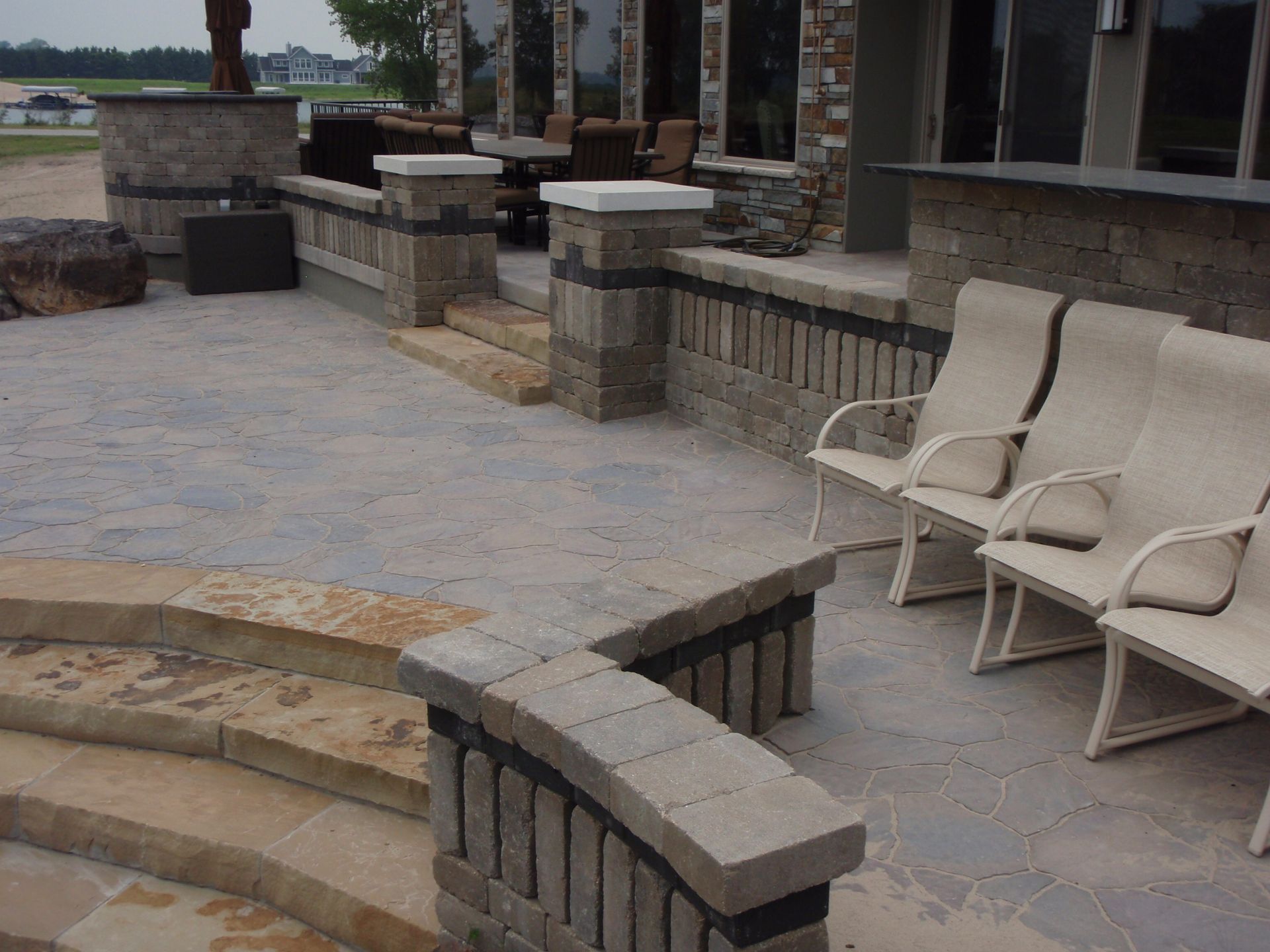 Stone patio with curved steps and beige lounge chairs beside a brick building.