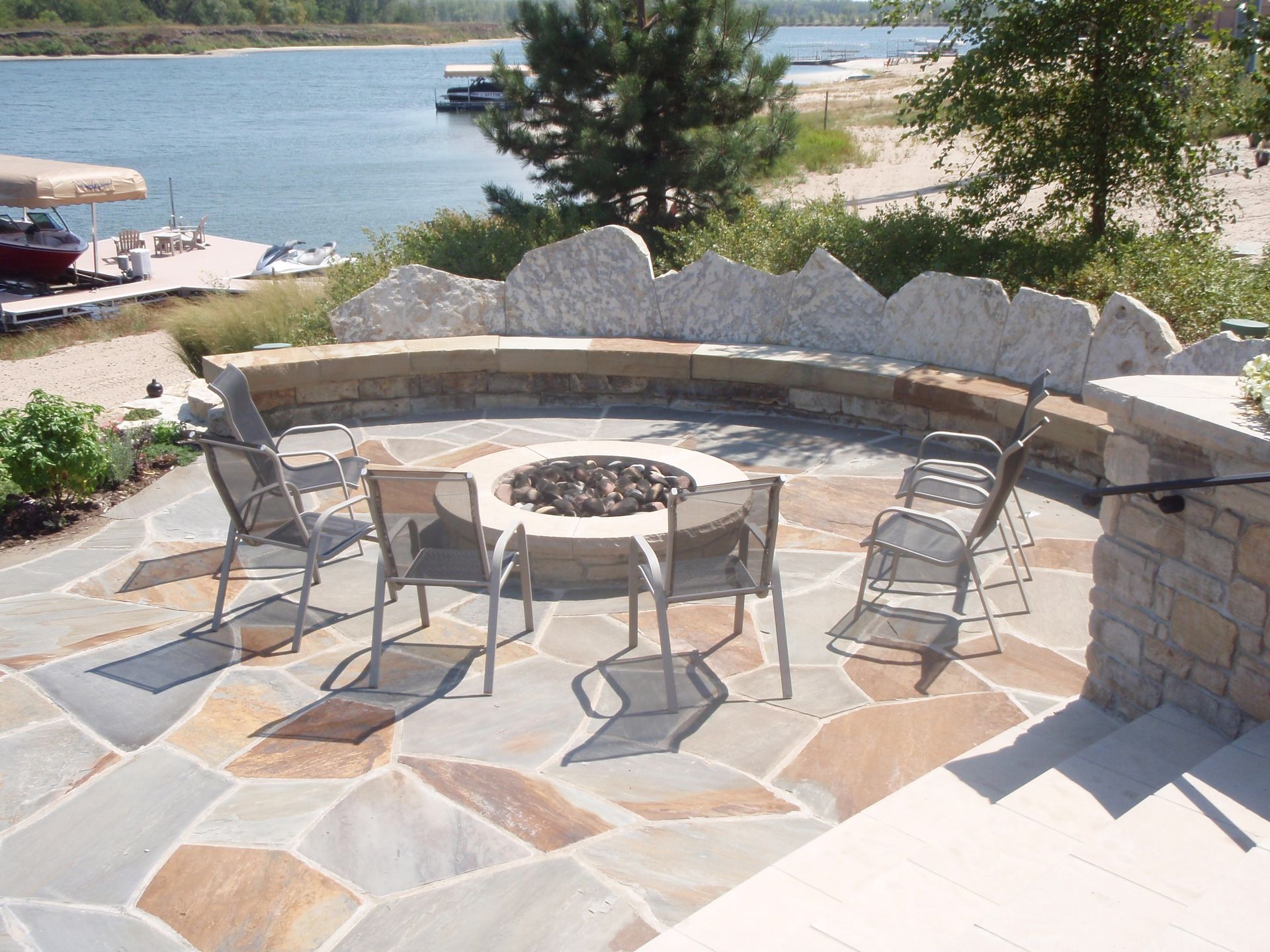 Stone patio with fire pit and metal chairs overlooking a lake and sandy shore