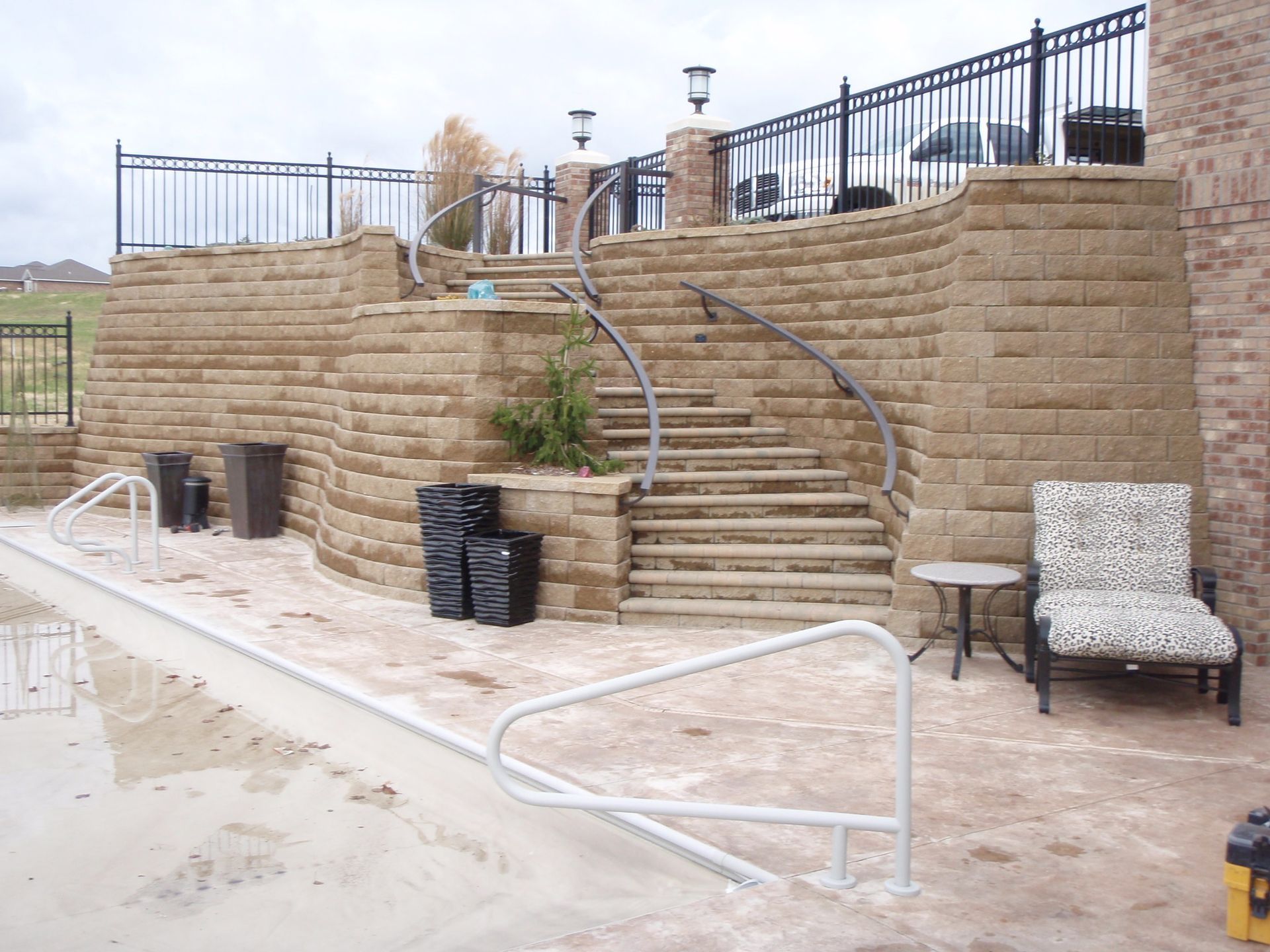 Stone backyard patio with central stairs, black metal railing, and outdoor seating near a poolside walkway