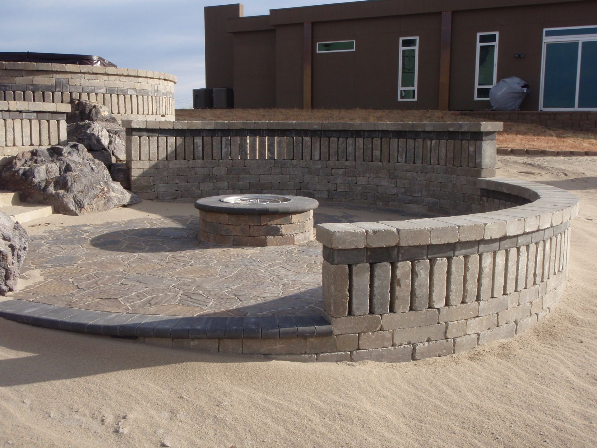 Circular stone seating around a fire pit in a sandy courtyard beside brick buildings