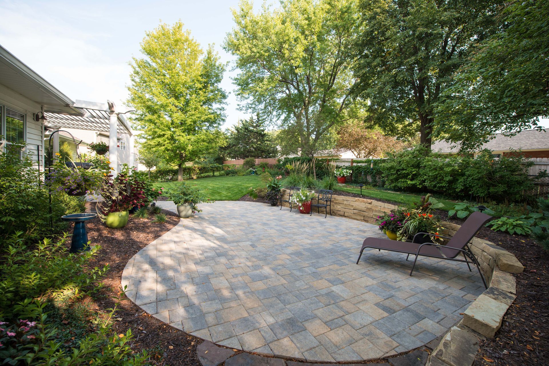 Backyard patio with stone pavers, lounge chair, and landscaped garden surrounded by trees and shrubs