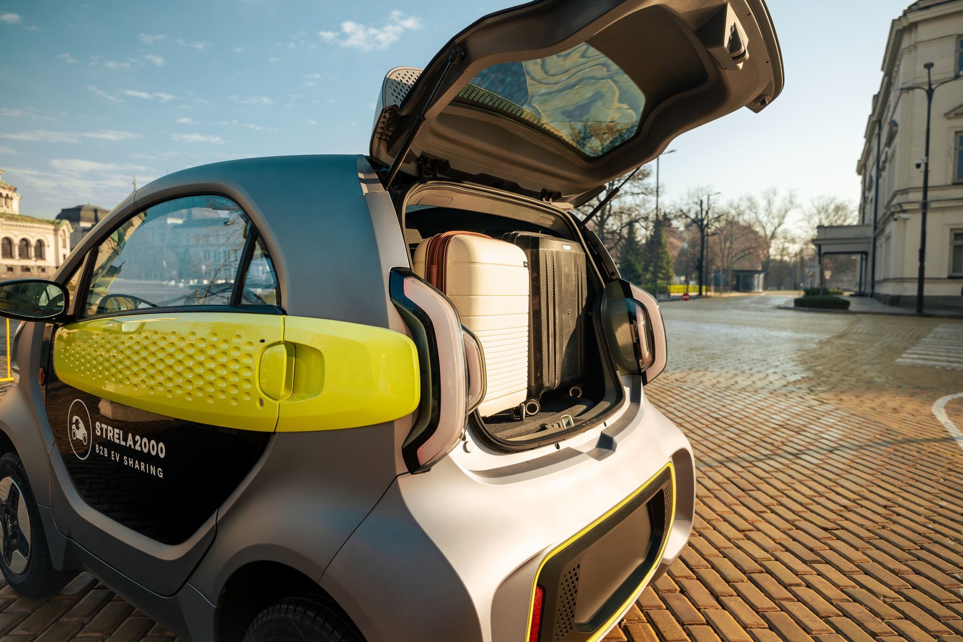 Small electric car with open rear hatch on a sunny cobblestone street near buildings