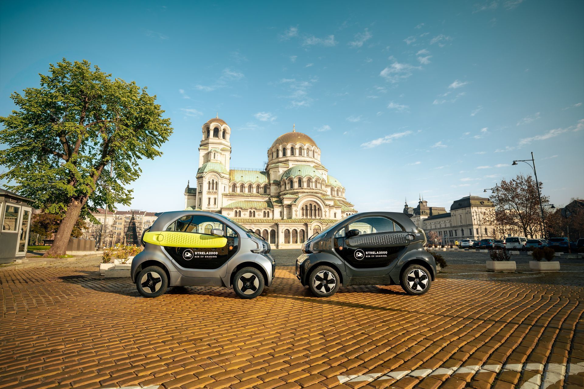 Two small electric cars parked on a cobblestone square in front of a cathedral under a blue sky