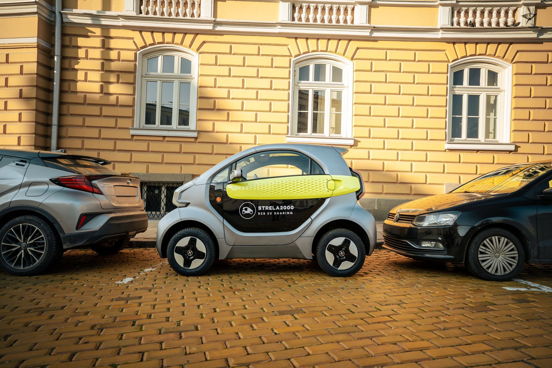 Three parked cars, including a small white Smart car, lined up on a cobblestone street by a stone building.