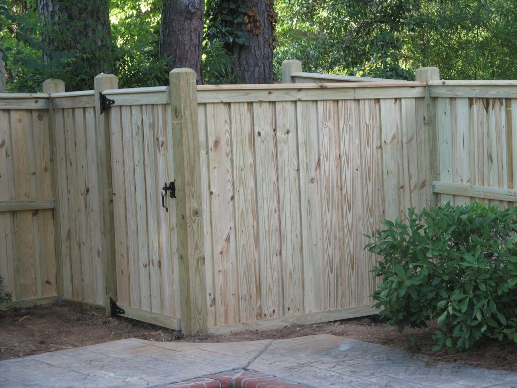 Wooden privacy fence with a gate beside a paved patio and shrubs in a backyard