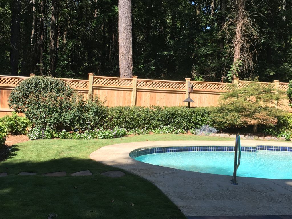 Backyard pool with turquoise water, stone deck, shrubs, and a wooden fence in the background