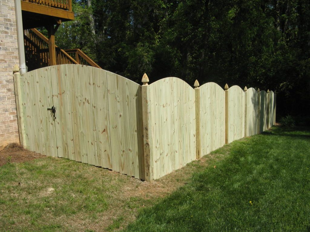 Light wood privacy fence with scalloped top in a grassy yard beside a house and trees