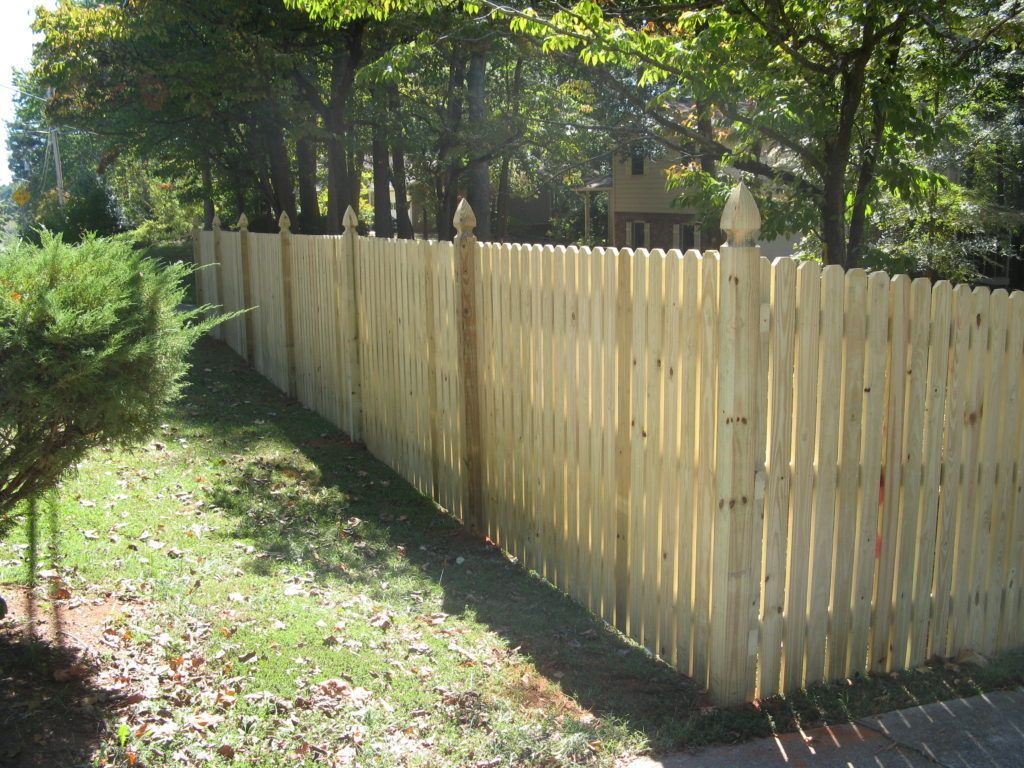 Light tan wooden picket fence along a grassy yard with trees in the background
