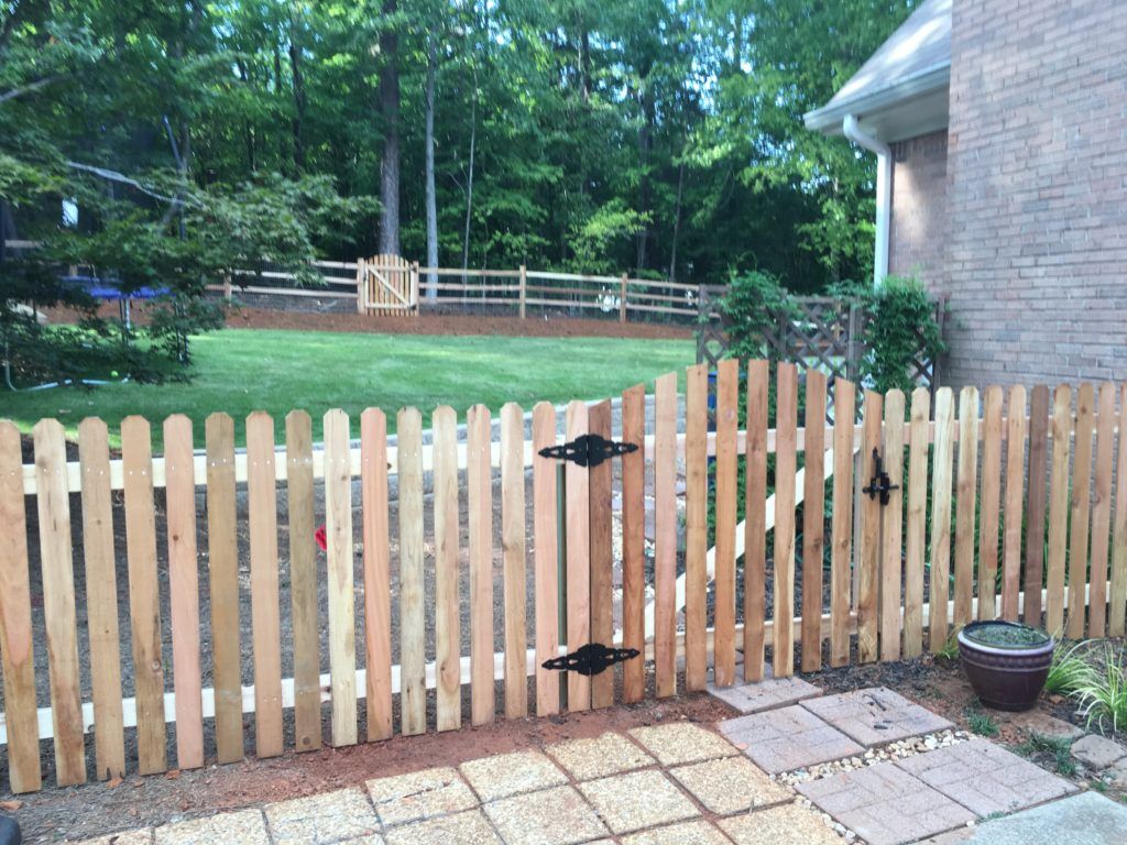 Wooden backyard gate and fence beside a brick house, with trees and lawn in the background.