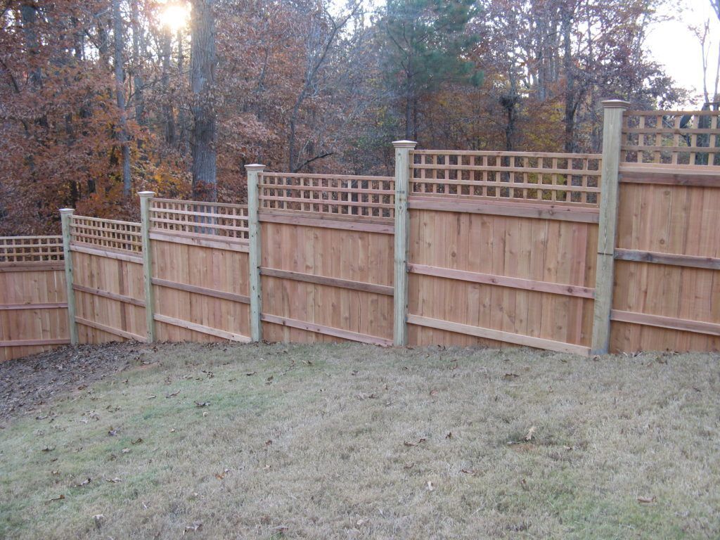 Wooden privacy fence with lattice panels along a grassy yard, backed by bare trees in autumn.