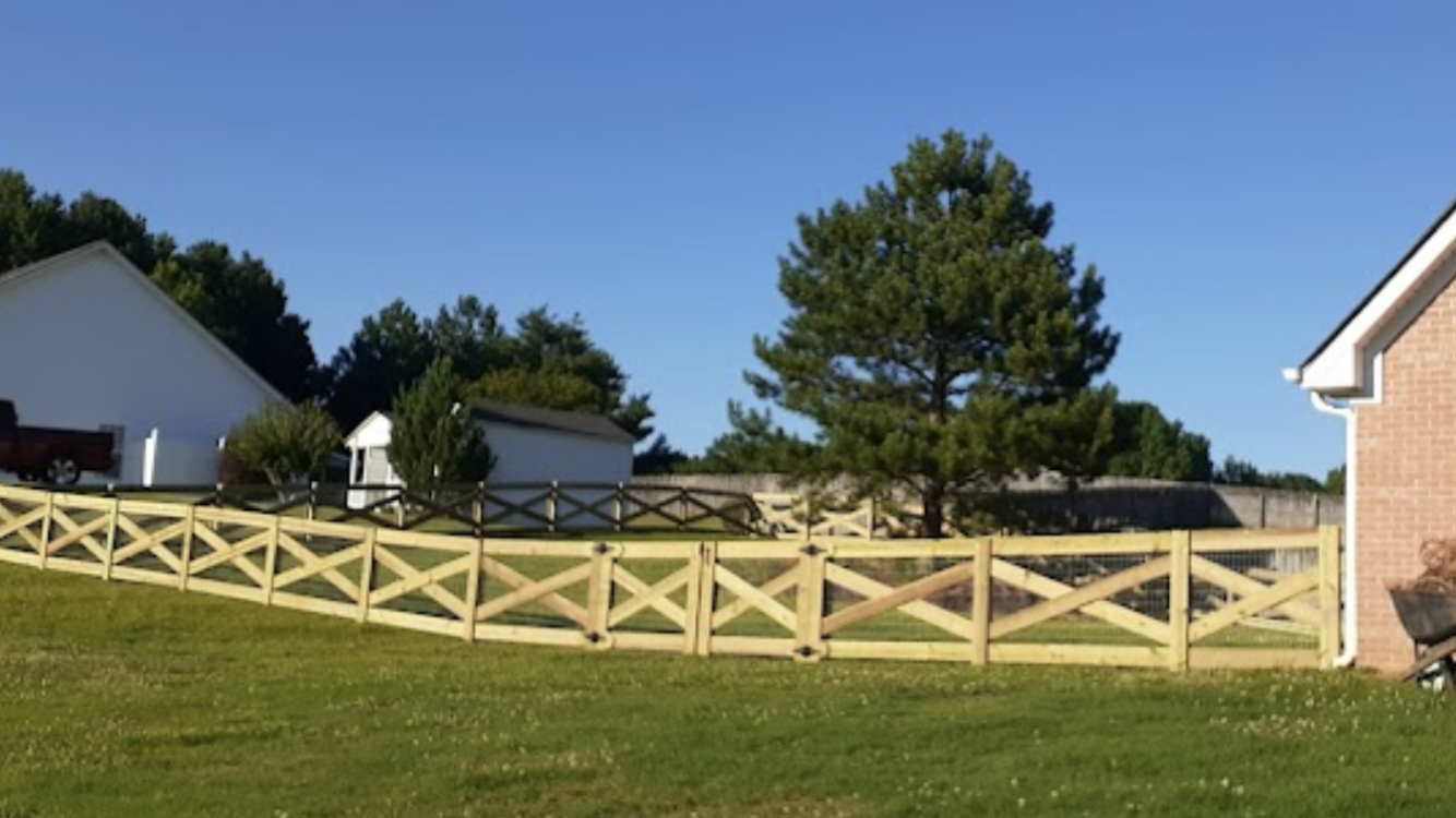 Wooden crossbuck fence in a grassy yard with houses and trees under a clear blue sky