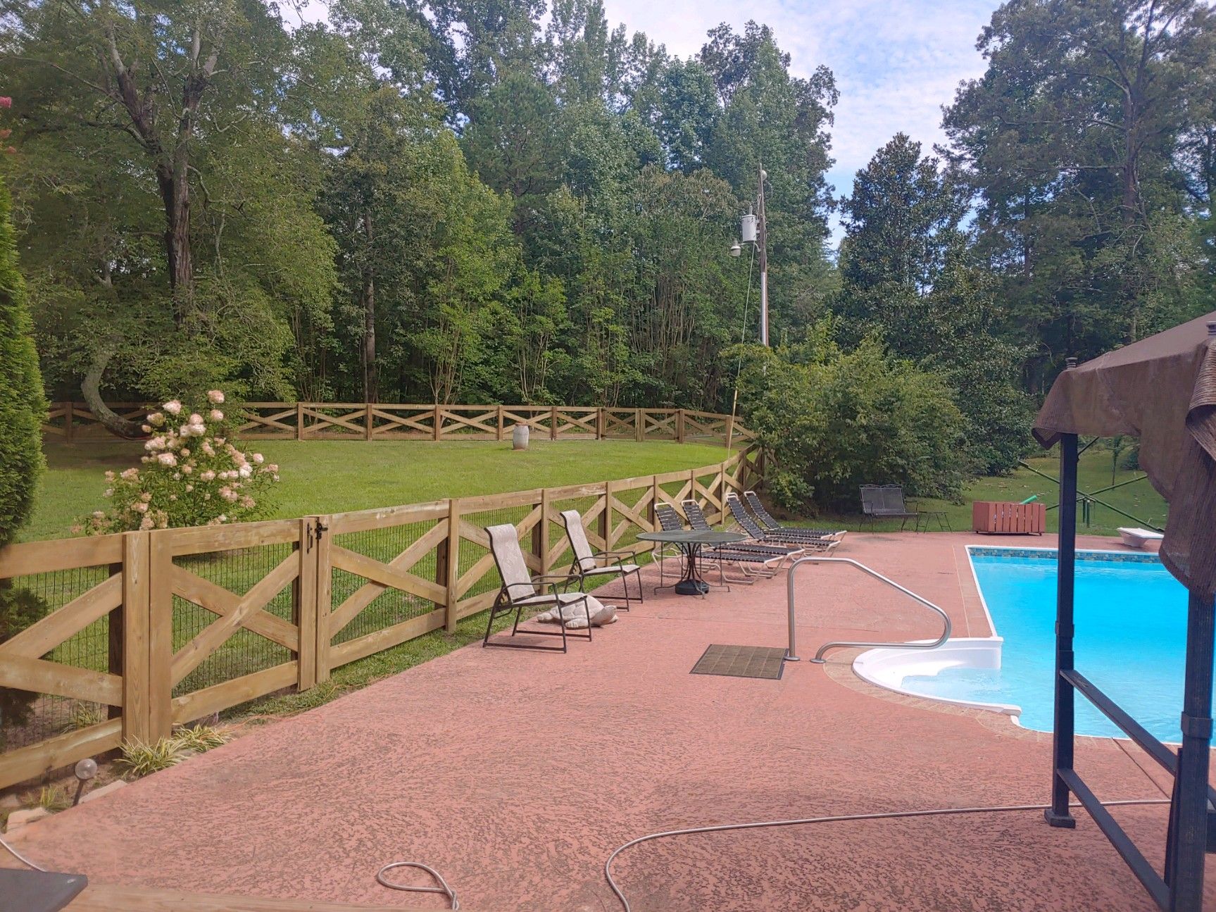 Outdoor pool deck with lounge chairs, wooden fence, and trees beyond the water