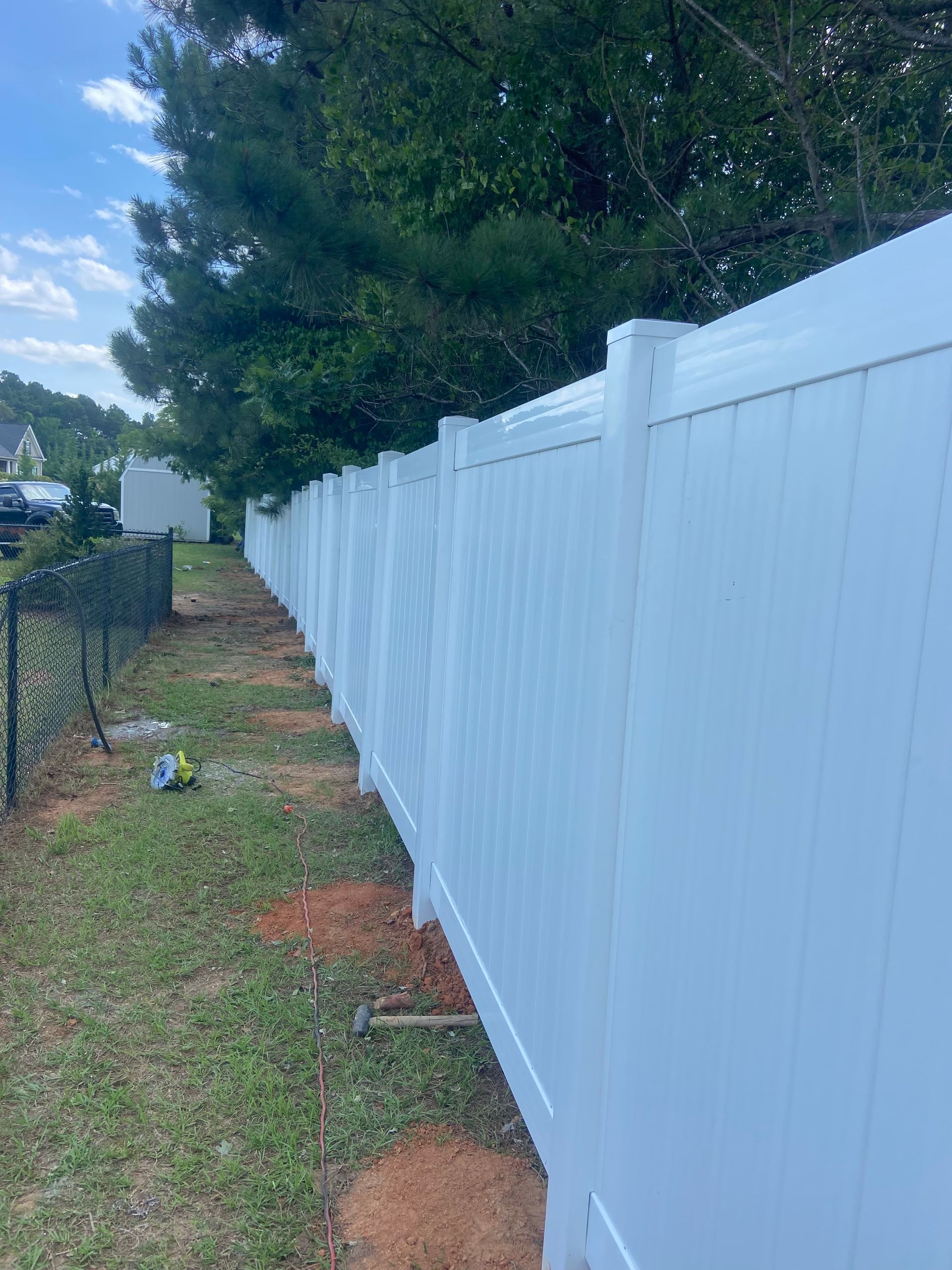 Long white vinyl fence along a grassy yard beside a chain-link fence and tall trees under a cloudy sky