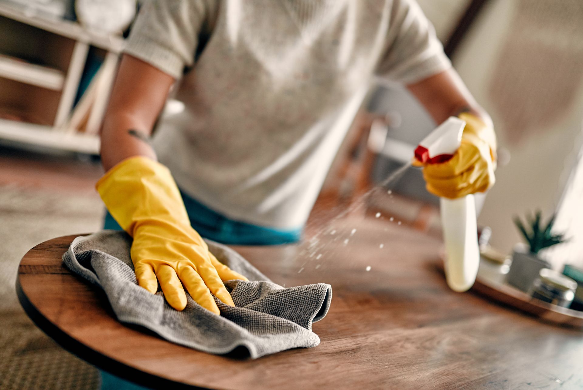 Person wearing yellow gloves spraying cleaner on a wooden table and wiping it with a cloth.