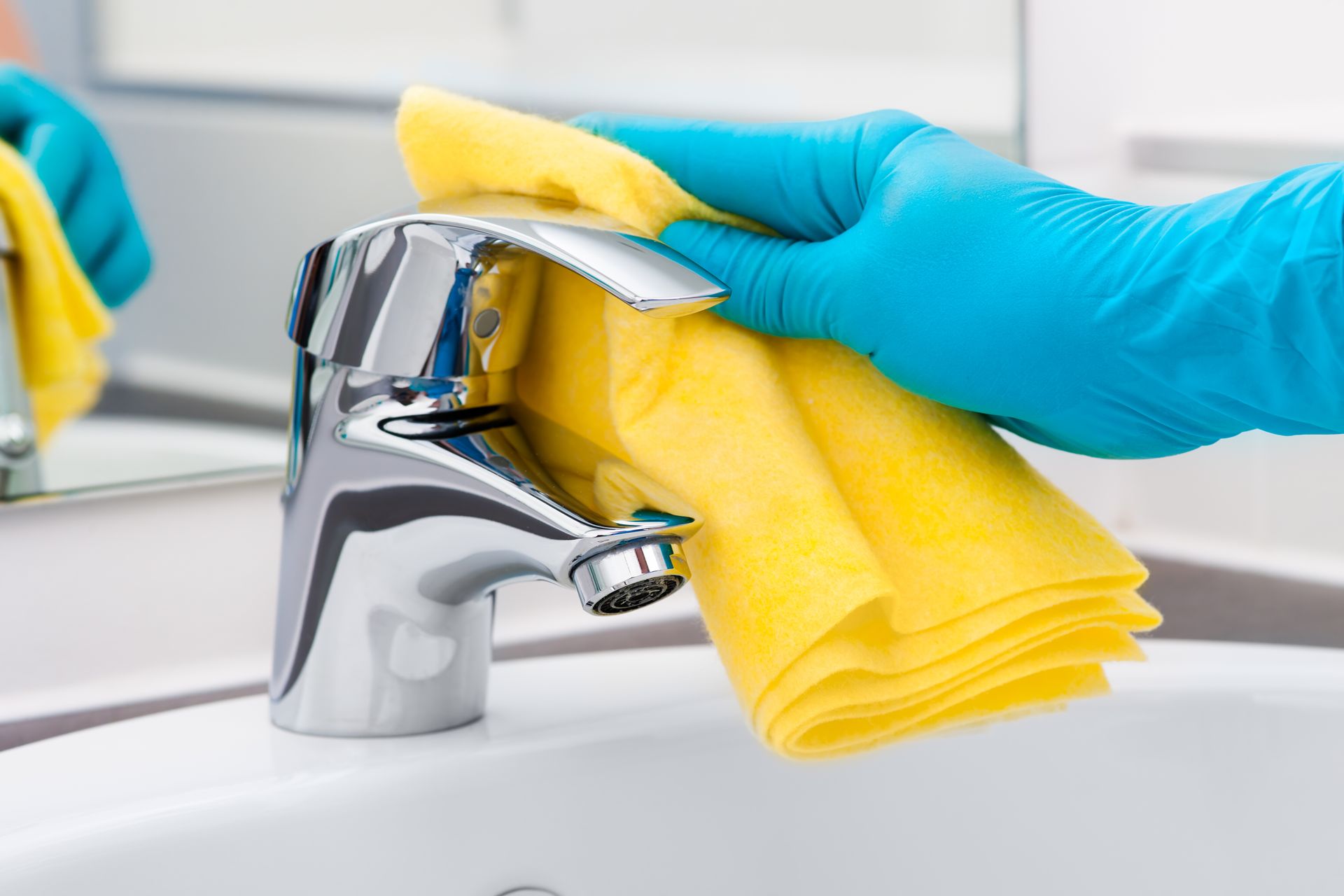A hand in a blue glove wipes a chrome faucet with a yellow cloth over a white sink.
