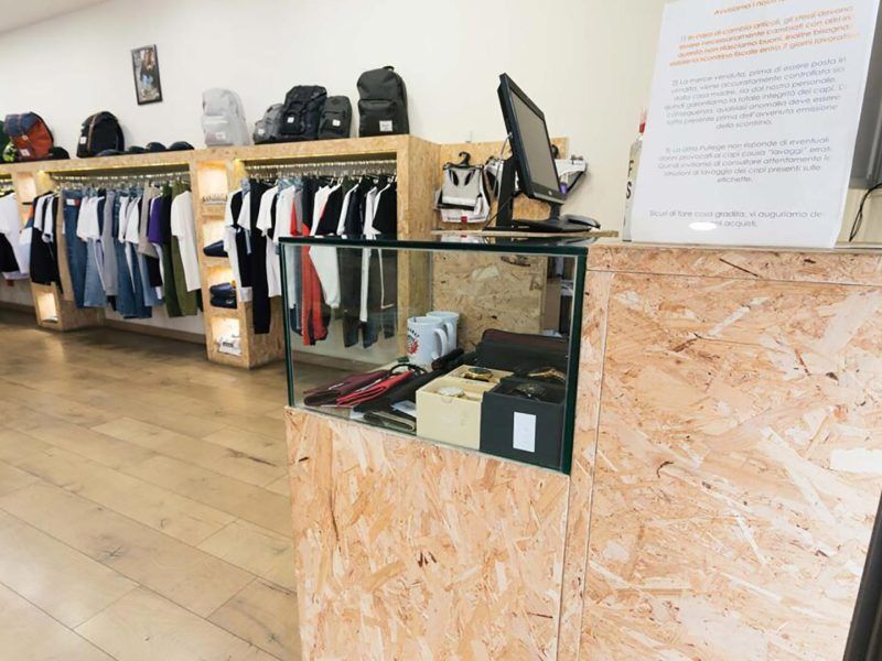 Interno di un negozio di abbigliamento con scaffali pieni di vestiti e una vetrina su un pavimento in legno.
