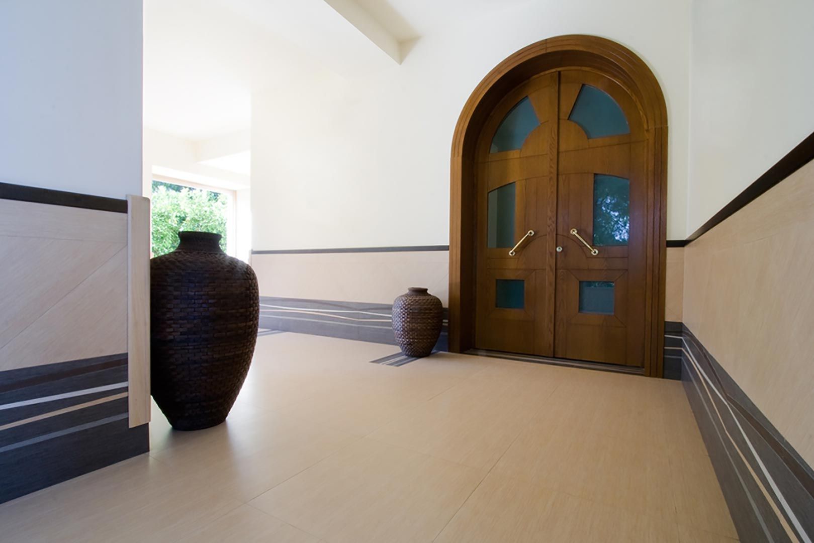Luminoso corridoio con doppia porta ad arco in legno, pavimento beige e grande vaso scuro accanto a una finestra soleggiata.