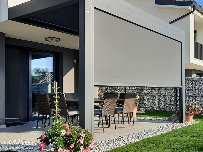 Patio coperto con set da pranzo all'aperto, fiori in vaso e muro decorativo in pietra accanto a una casa moderna.