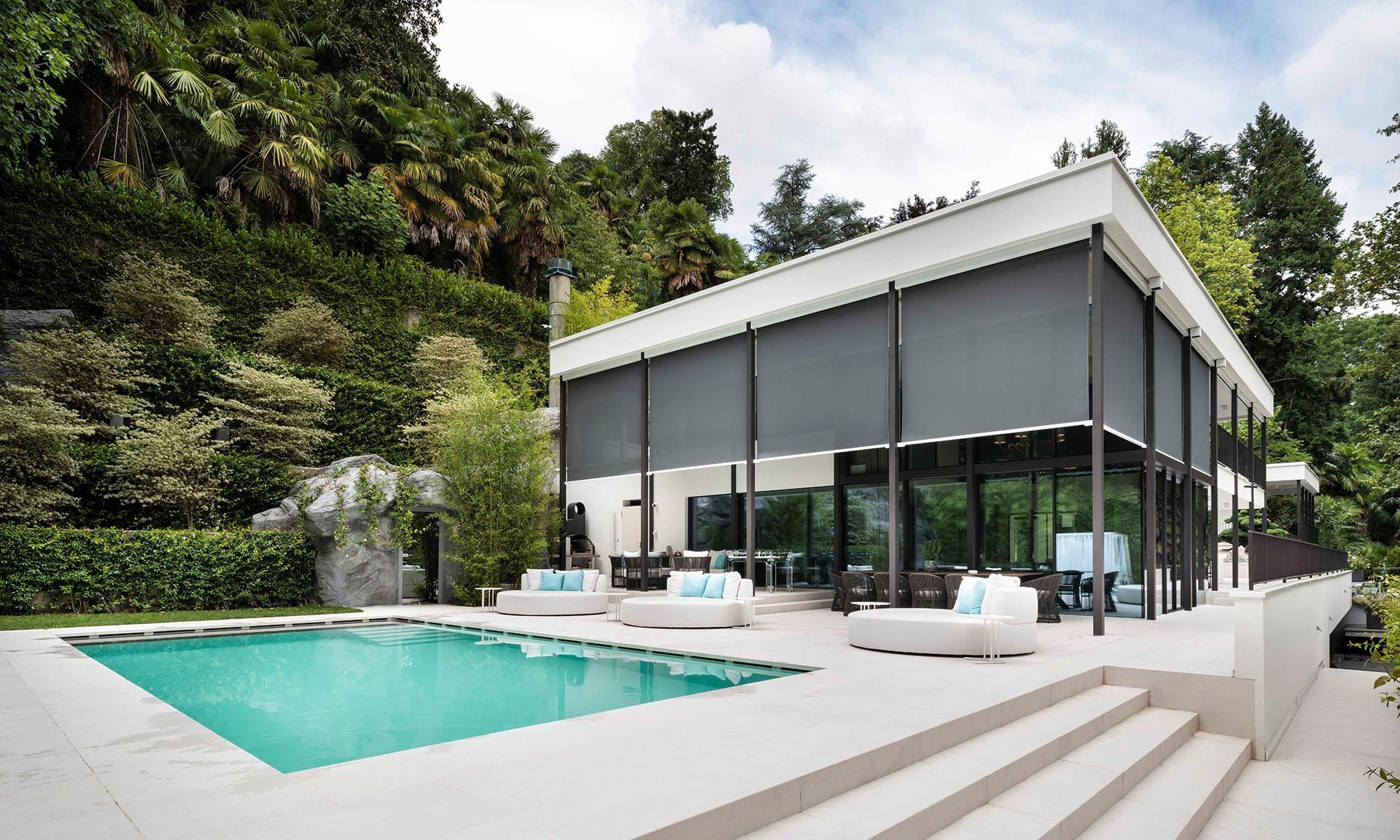 Casa moderna bianca con piscina turchese e lettini prendisole immersi nel verde lussureggiante.