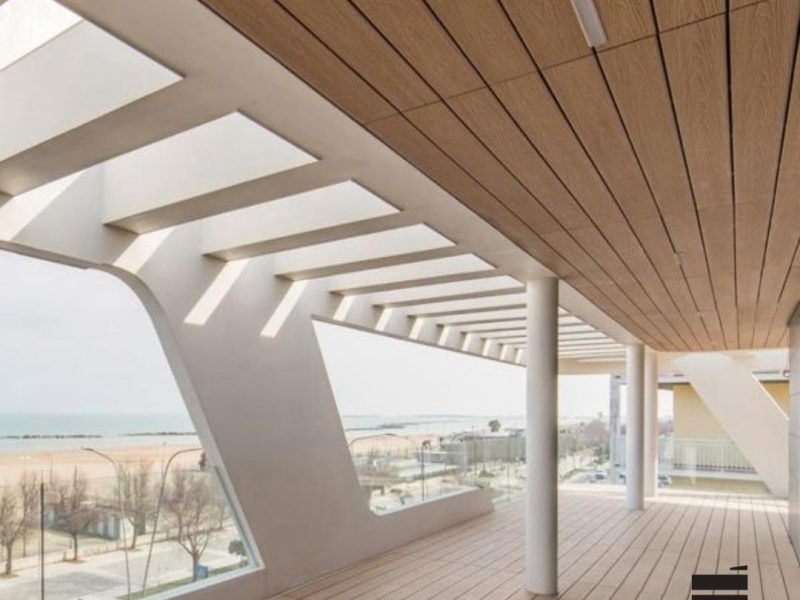 Moderna passerella coperta con colonne bianche, soffitto in legno e vista sul mare.