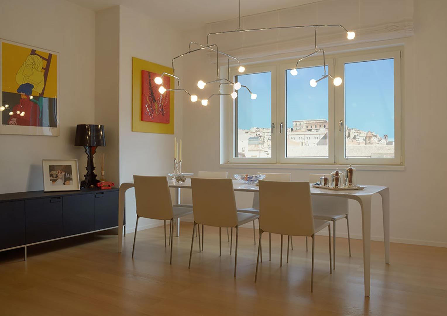 Luminosa sala da pranzo con tavolo e sedie bianchi, lampade a sospensione e ampie finestre che si affacciano sugli edifici della città.
