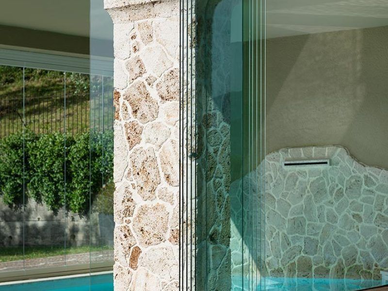 Una porta a vetri accanto a una colonna di pietra riflette un paesaggio verdeggiante e rivela all'interno una zona piscina piastrellata.